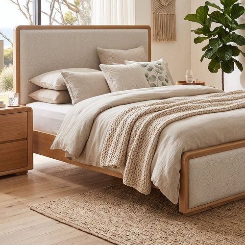 A modern bedroom featuring a light wood and upholstered bed frame, styled with neutral beige bedding, a matching wooden nightstand, a wooden chest of drawers, and a large potted plant in front of a large window overlooking trees and the ocean.