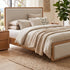 A modern bedroom featuring a light wood and upholstered bed frame, styled with neutral beige bedding, a matching wooden nightstand, a wooden chest of drawers, and a large potted plant in front of a large window overlooking trees and the ocean.